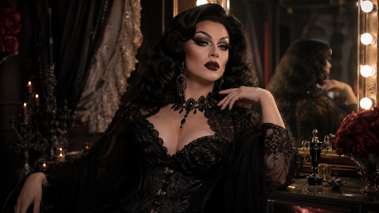 Drag queen avec look dark glam, maquillage intense, perruque volumineuse, tenue noire brillante et bijoux imposants dans une loge de spectacle.