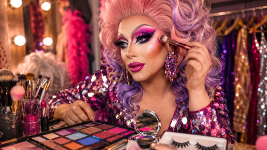 Maquillage drag queen débutant avec pinceaux, fards colorés, faux cils, rouge à lèvres et accessoires glamour sur une table de préparation.